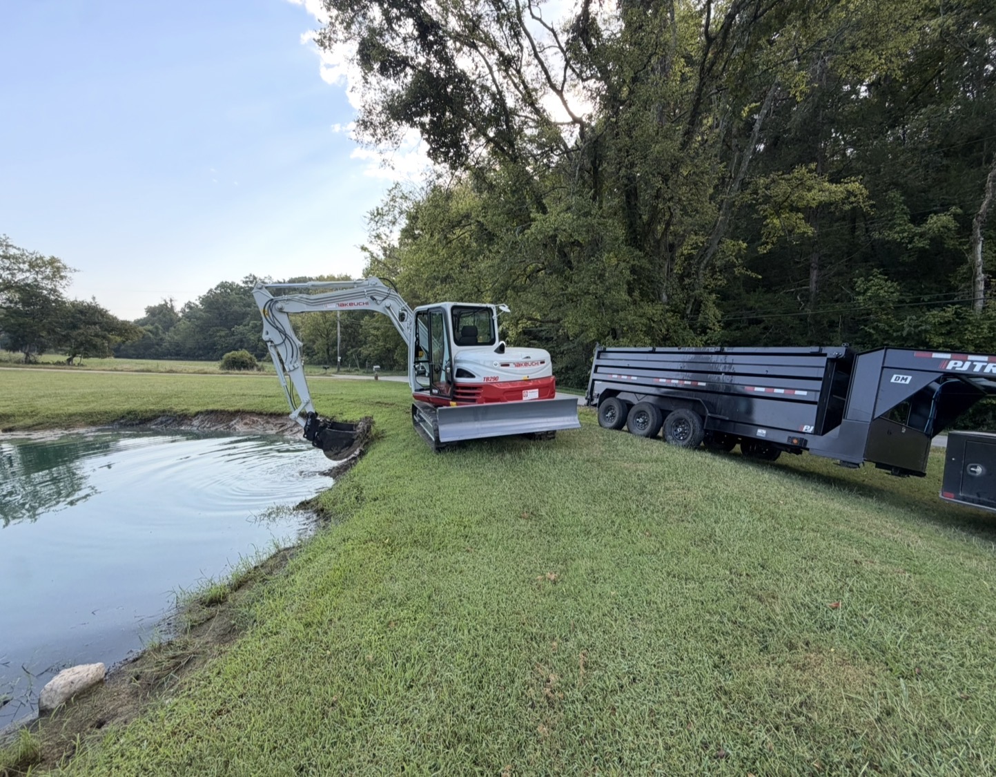 Excavator working along the bank of a pond with a dump trailer nearby